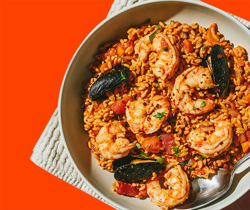 A bowl of seafood paella with shrimp, mussels, and saffron-infused rice, garnished with herbs. The bowl rests on a textured white cloth with an orange background.