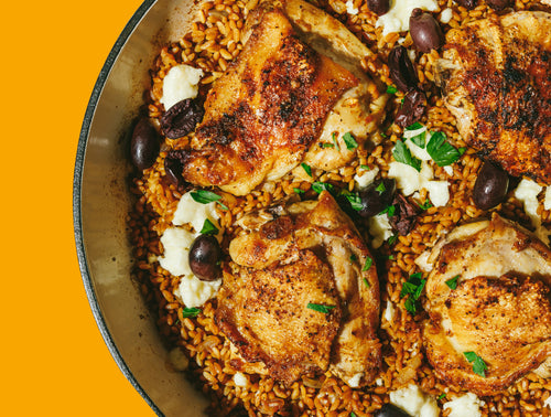 Pan with seasoned baked chicken thighs, brown rice, olives, and scattered cheese, garnished with parsley, against a bright orange background.