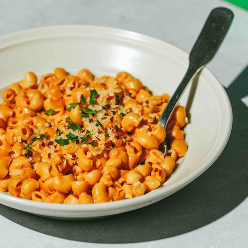 A bowl of pasta in tomato sauce, topped with grated cheese and chopped herbs, sits on a light-colored surface. A fork is placed in the bowl, casting a shadow. The dish is brightly lit, highlighting the textures and colors.