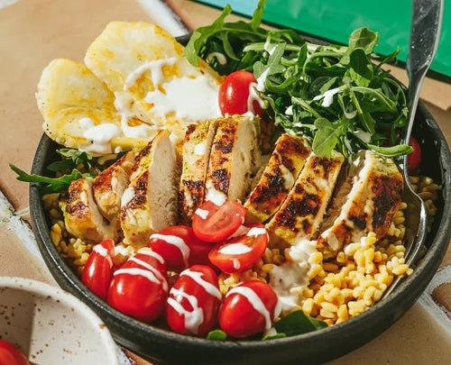 A bowl filled with grilled chicken slices, cherry tomatoes, arugula, pineapple slices, and grains, drizzled with white dressing. A fork is placed in the bowl. The meal is displayed on a tiled surface.