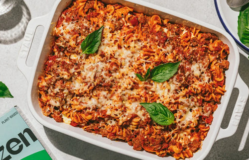 A white baking dish filled with baked pasta topped with melted cheese, ground meat, and tomato sauce. Fresh basil leaves garnish the top. A cookbook corner and a glass of water are partly visible nearby.