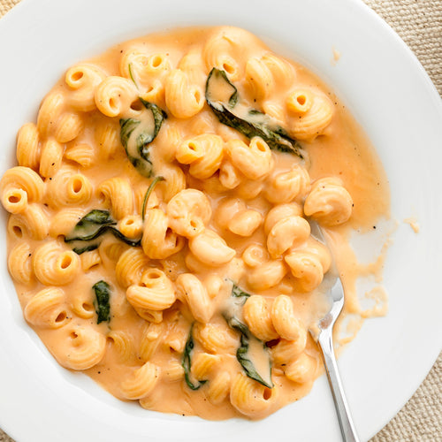 A white plate of Lowcarb, High Protein, Plant Based Pasta (SYS) by Kaizen Food Company in a rich orange sauce with fresh spinach. A fork rests slightly immersed in the pasta on a woven beige placemat.